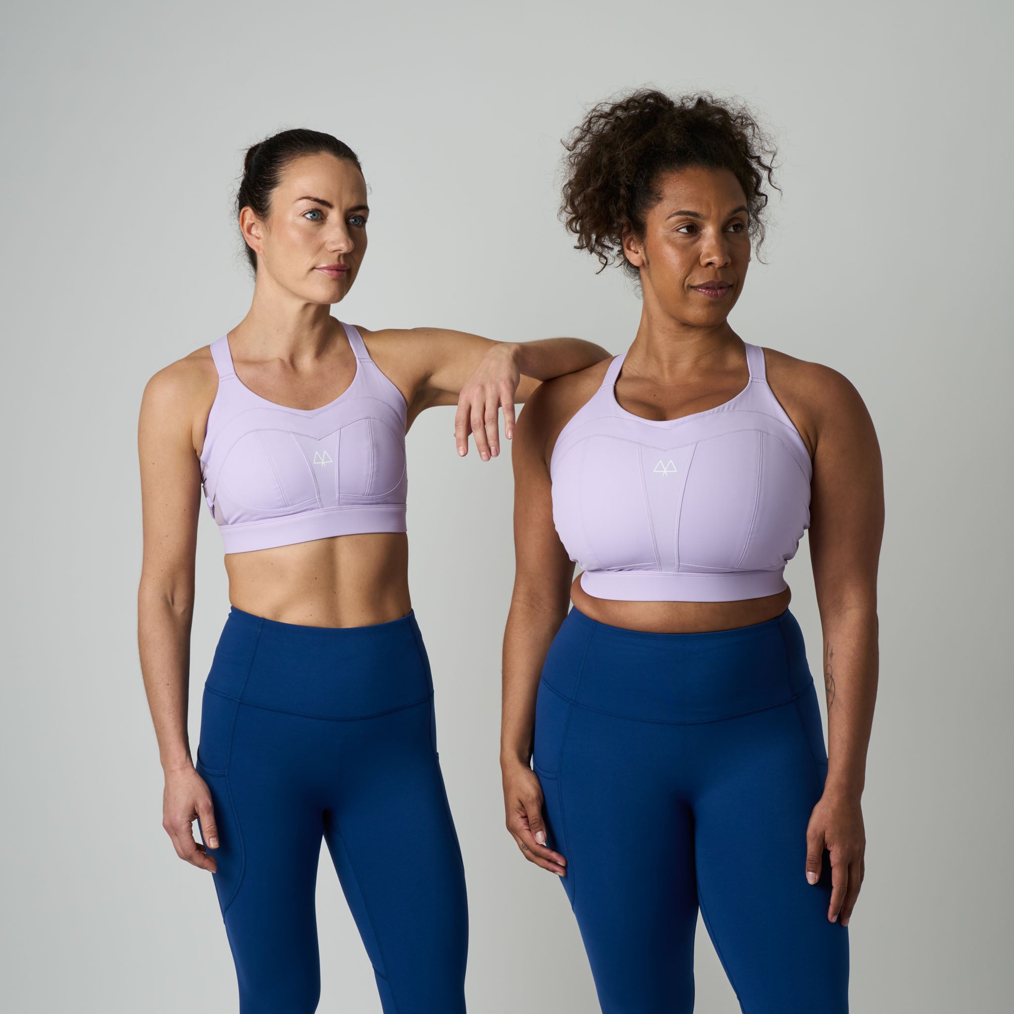 Two women wearing matching purple sports bras and blue leggings against a plain background