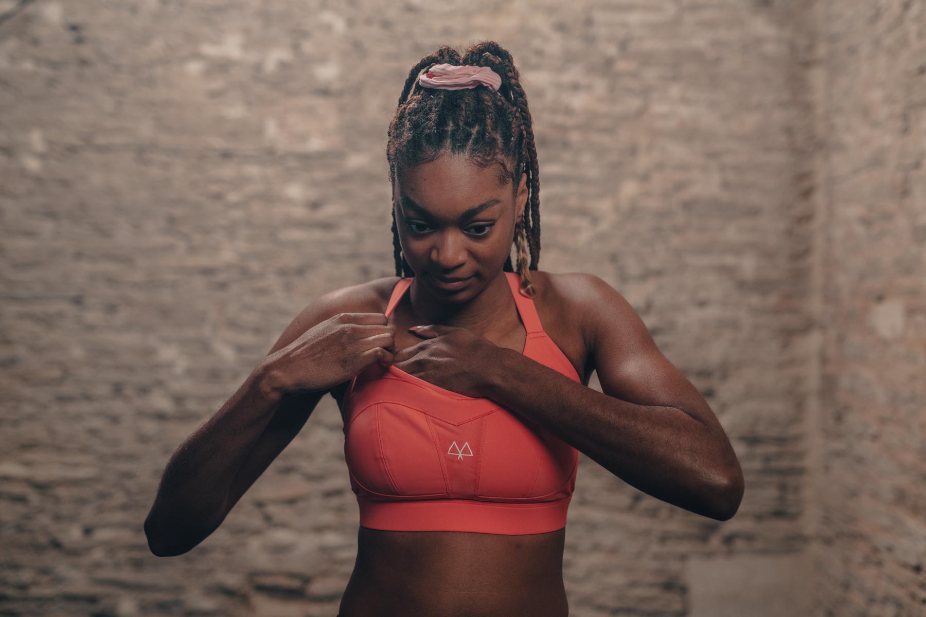 Girl adujsting her bright orange sports bra whilst standing in front of a neutral brick wall